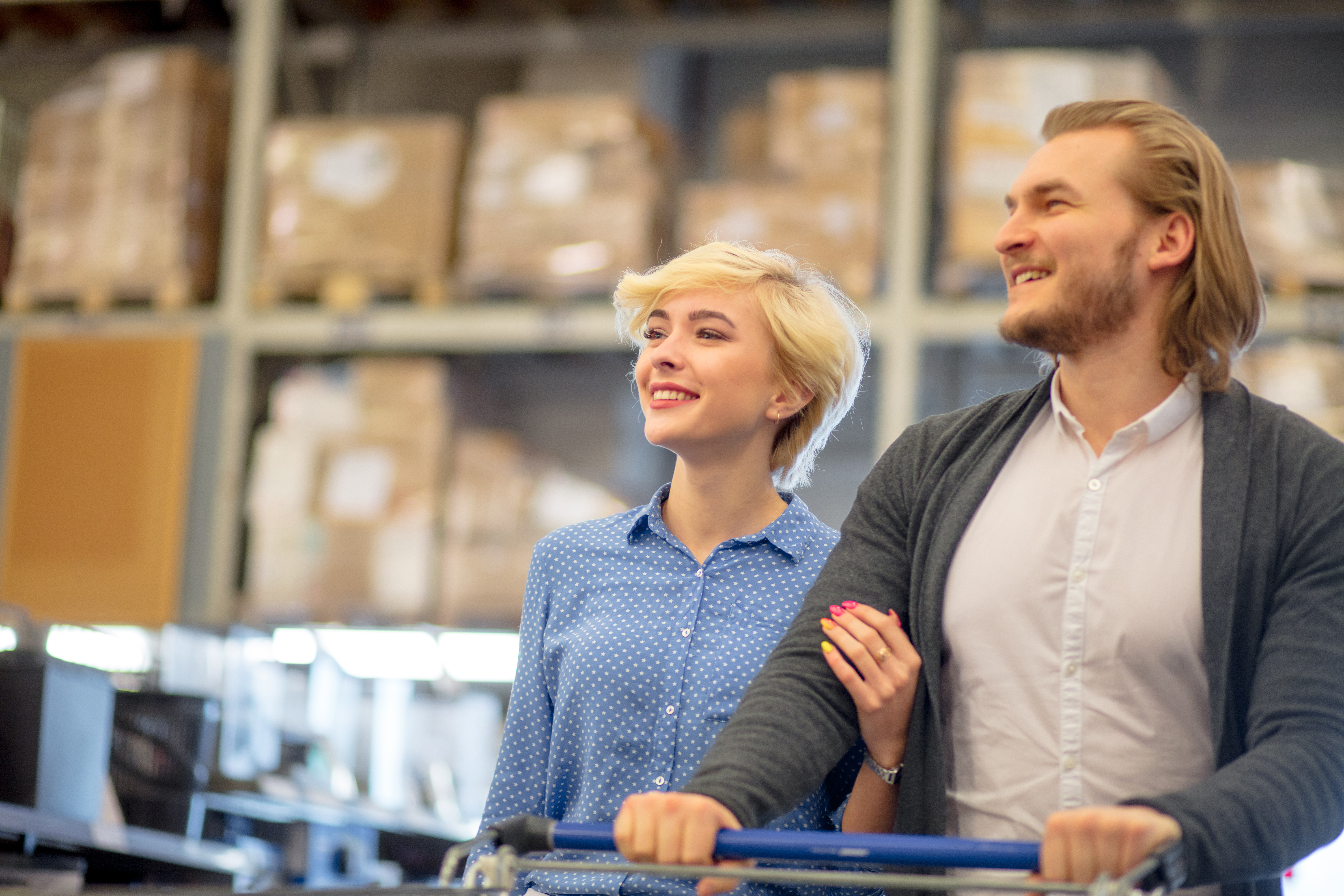 Happy married couple choosing goods in supermarket. Adult husband in company with his pretty blonde wife pushing shopping cart on big shopping mall.