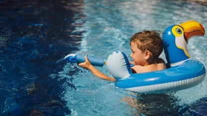 A boy with a joyful expression is floating in a swimming pool on a sunny day, using an swimming circle designed to look like a blue bird.