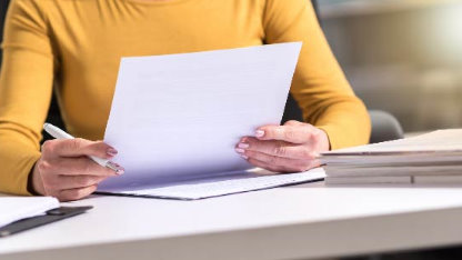 Buste d'une personne avec un pull jaune, assise à un bureau avec des dossiers dessus et tenant des papiers